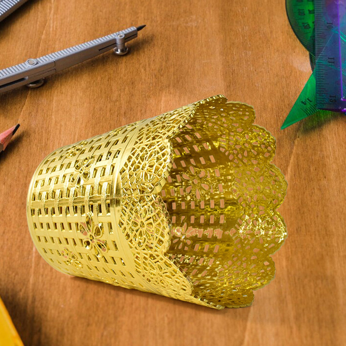 A gold plastic pen holder with a floral lace top rim lies on its side on a wooden surface, next to a ruler.