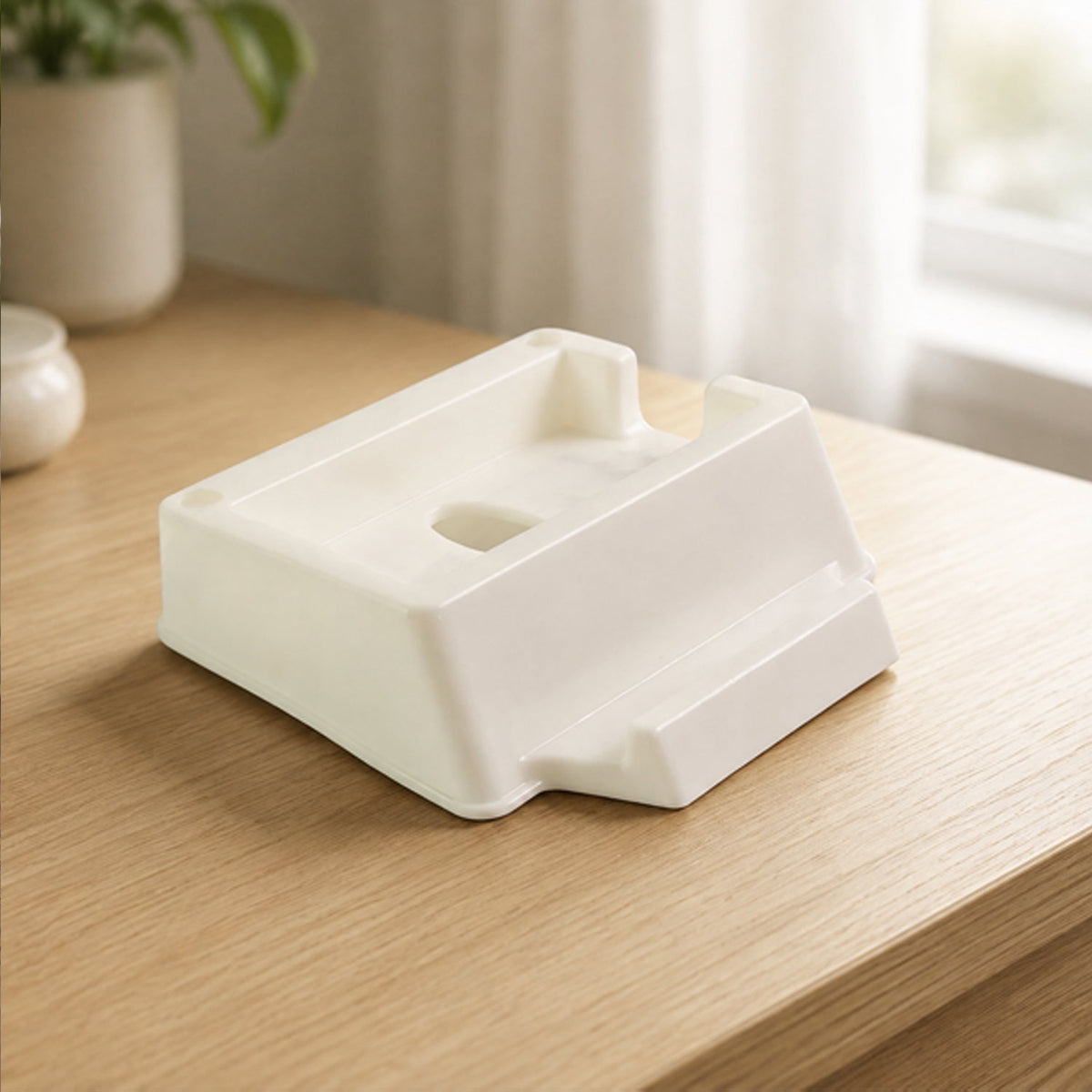 The cream plastic phone stand sits empty on a light wooden desk, with a window and plant in the background.