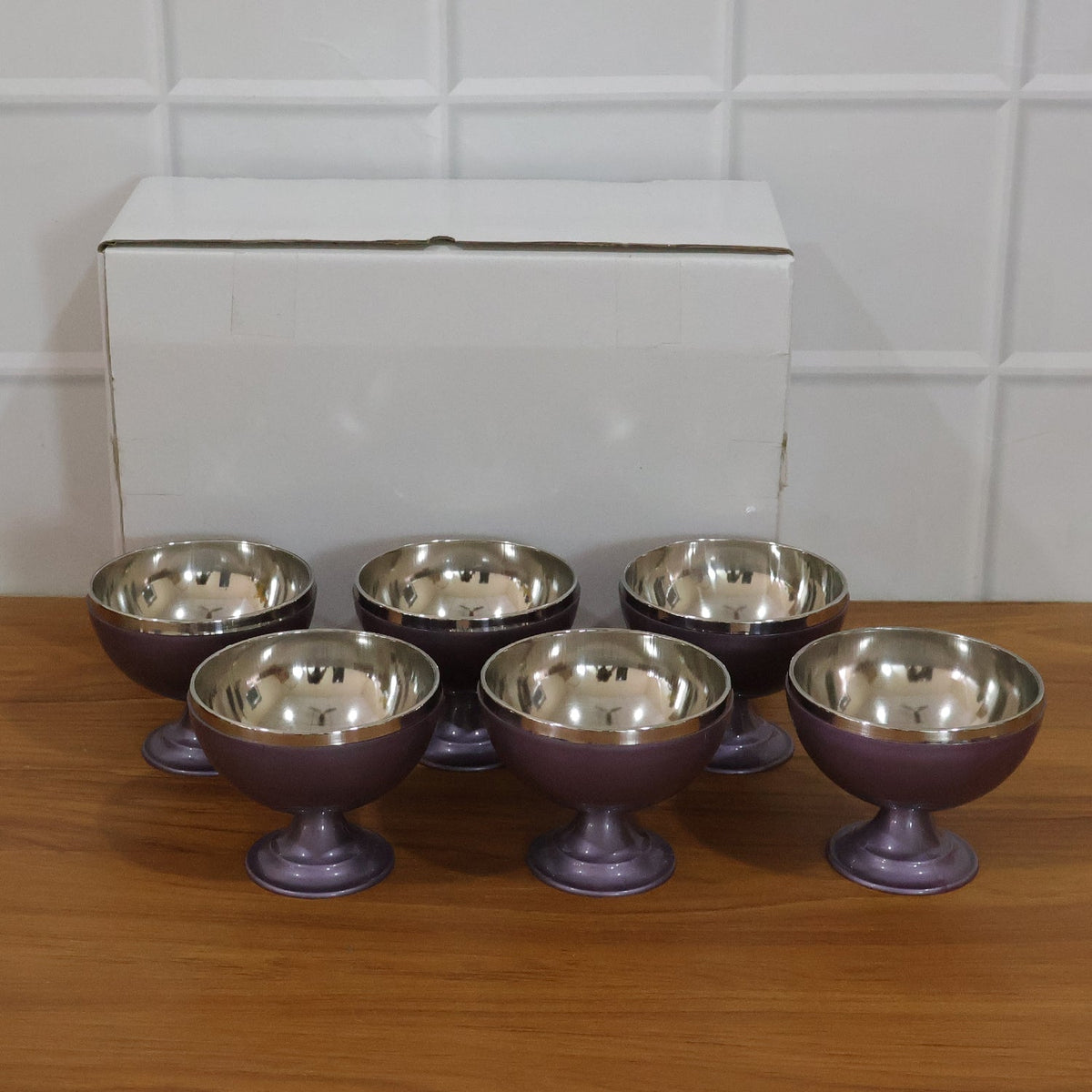 Six purple and silver dessert bowls are lined up on a wooden surface in front of a white box.