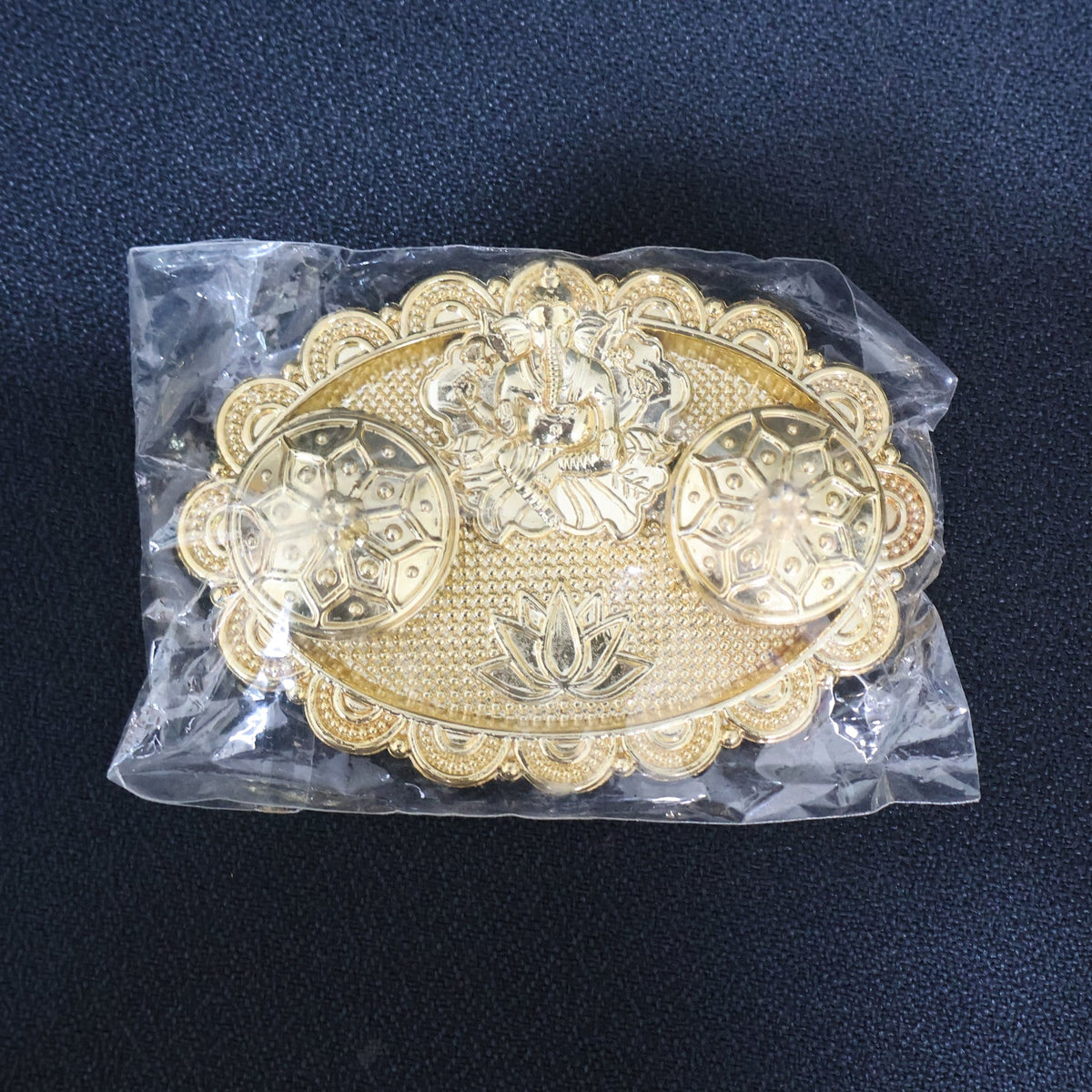 A gold-finished chopra with Ganesh and ornate containers, sealed inside a clear plastic bag on a dark background.