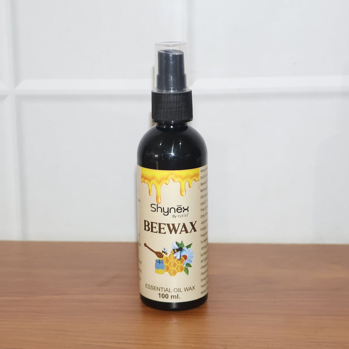 A black 100ml spray bottle of Shynex Beeswax essential oil wax stands on a wooden surface against a white tiled wall.
