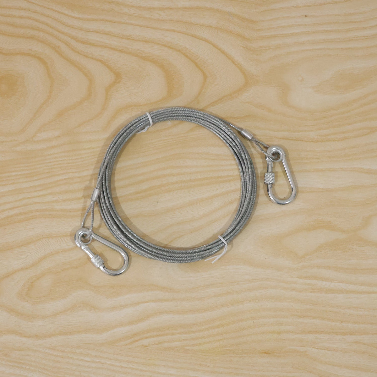 A coiled silver wire rope with two attached locking hooks on a light wooden background.