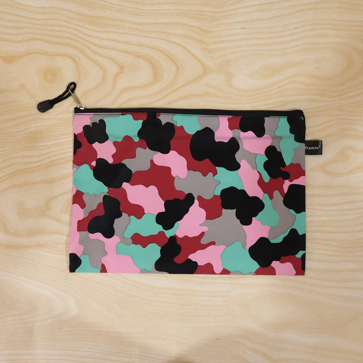 A flat, closed pouch with a pink, red, black, grey, and teal camouflage pattern, featuring a black zipper and pull tab, on a