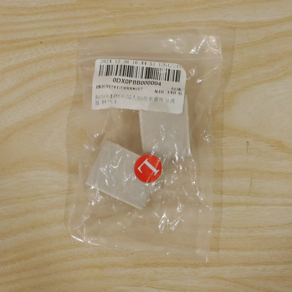 A clear plastic bag containing two beige elastic toe separators and a red circular sticker with the letter "L", lying on a