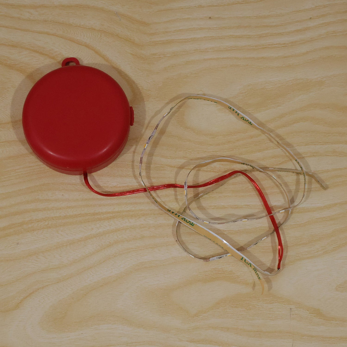 A red battery pack with a tangled LED strip light and red wire is placed on a light wooden surface.