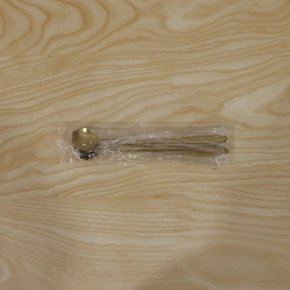 Three gold spoons are sealed in clear plastic packaging on a light wooden surface.