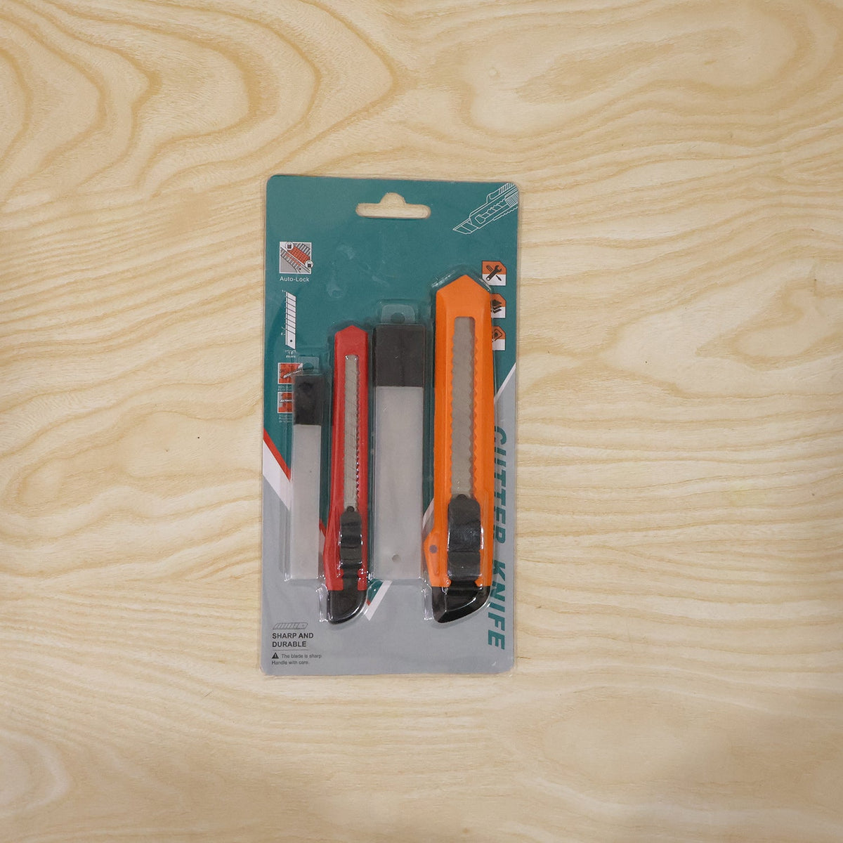 A packaged set of two utility knives, one red and one orange, with extra blades, on a light wooden background.