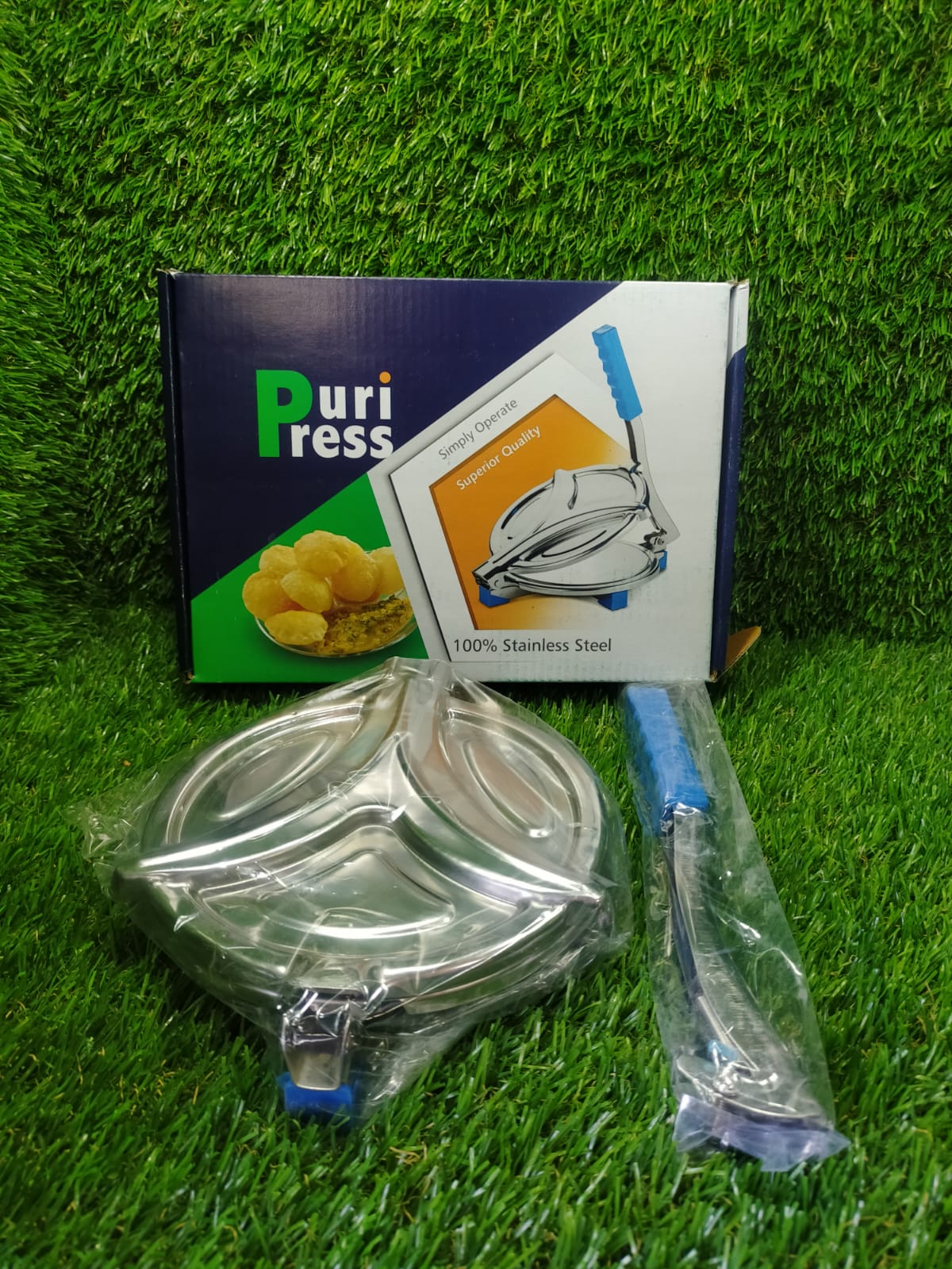 A packaged stainless steel puri press with blue handle is displayed on green artificial grass next to its box.