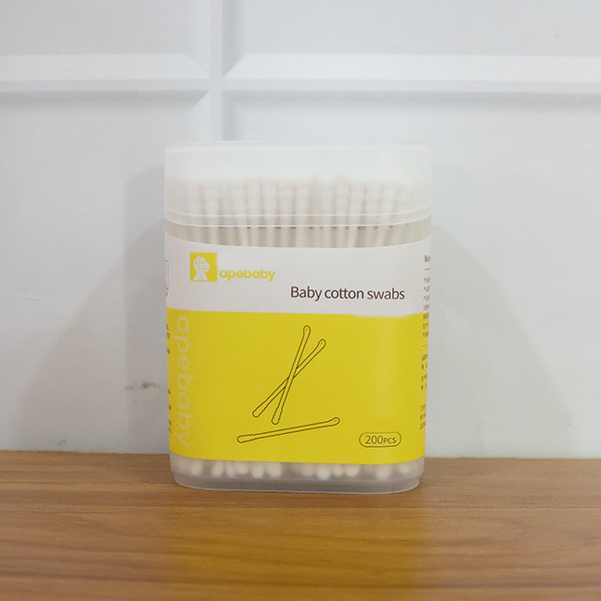 A clear plastic container of white baby cotton swabs with a yellow label, featuring product branding and a 200 pcs count, on