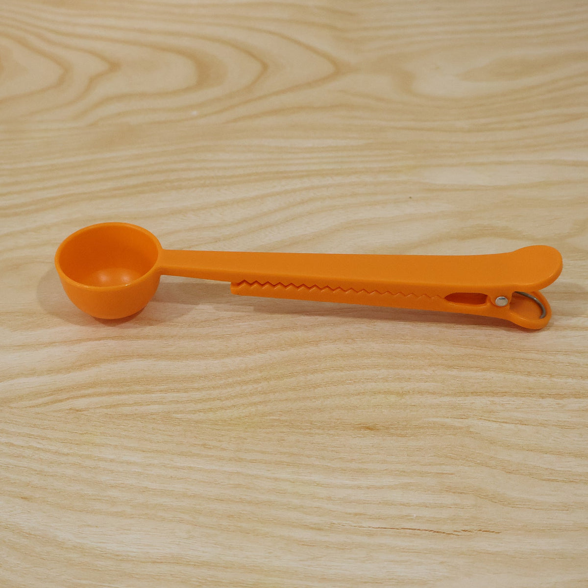 A simple orange plastic measuring spoon with a clip is positioned on a light brown wooden surface.