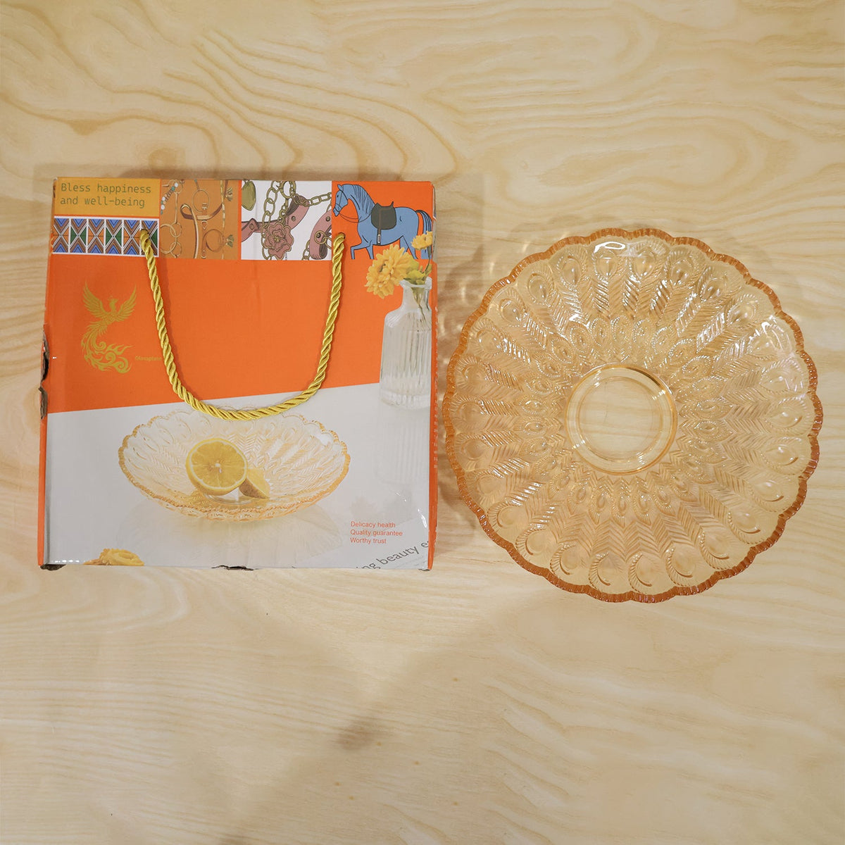 Amber glass platter with a feather design next to its orange gift box on a light wooden surface.
