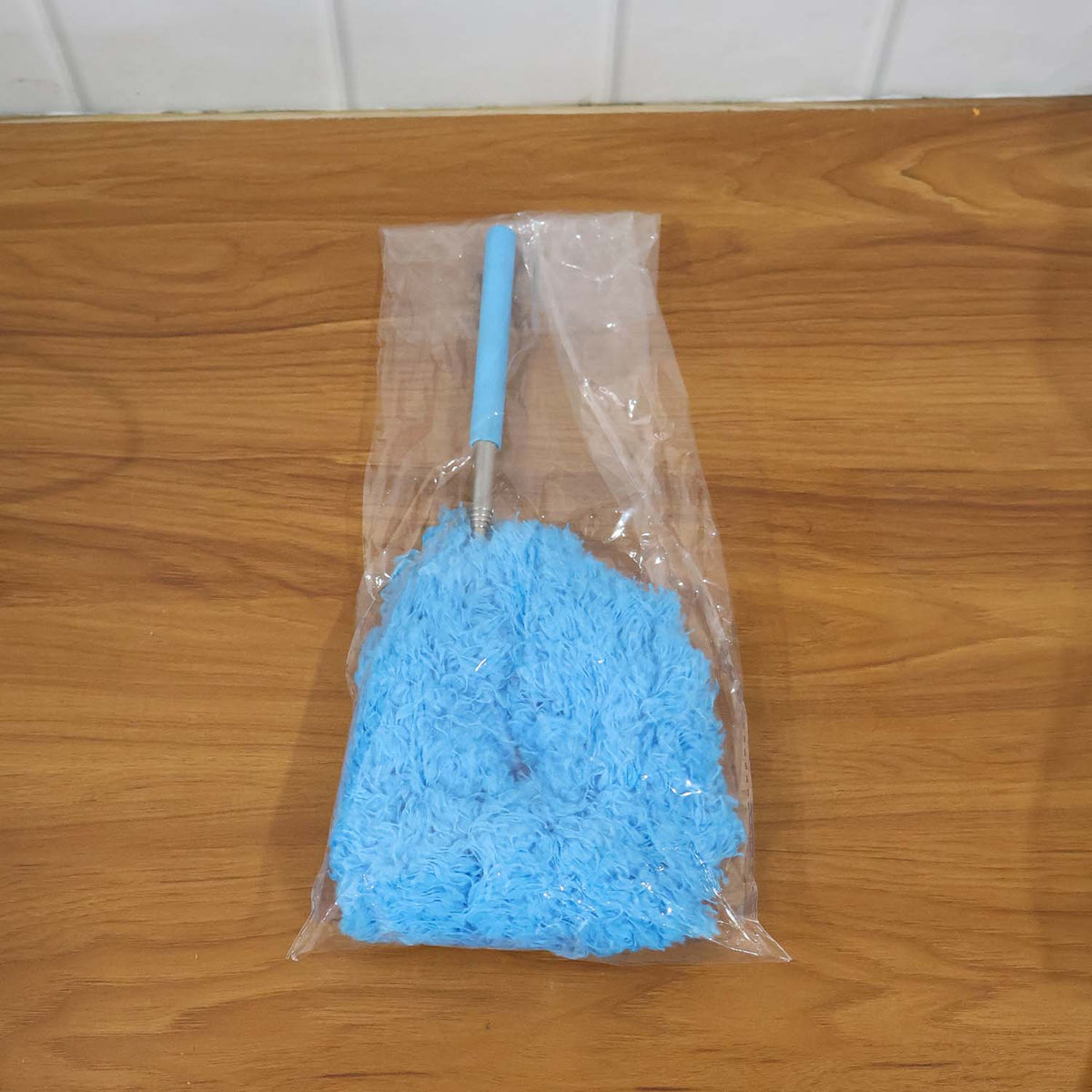 A blue microfiber duster with an extendable handle is packaged in clear plastic on a wooden surface.