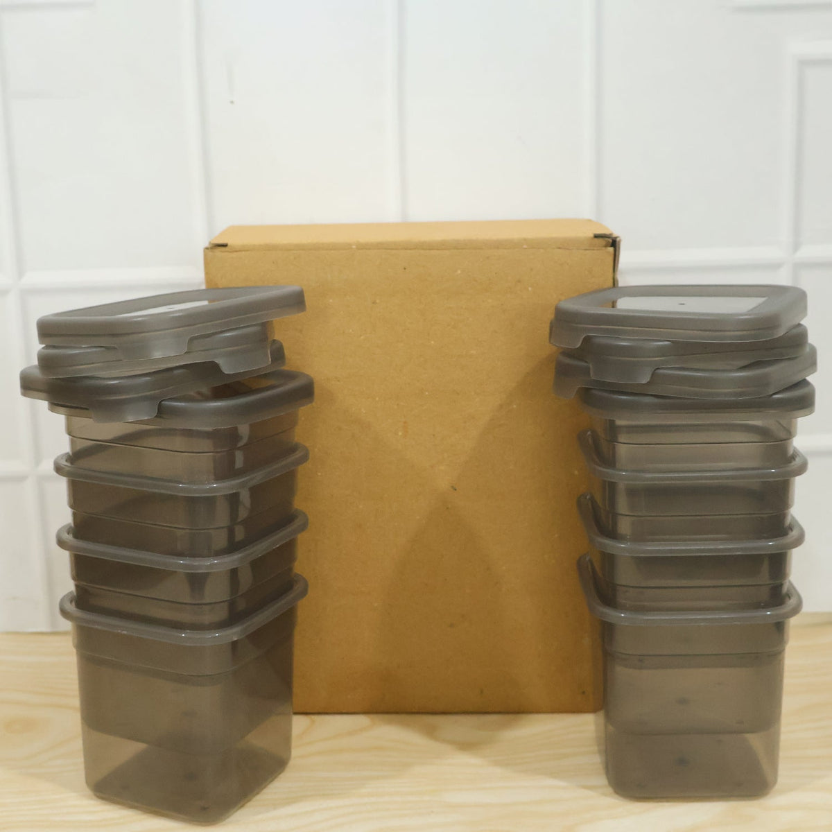 Two stacks of empty clear plastic food containers with gray lids, flanking a brown cardboard box on a wooden surface.