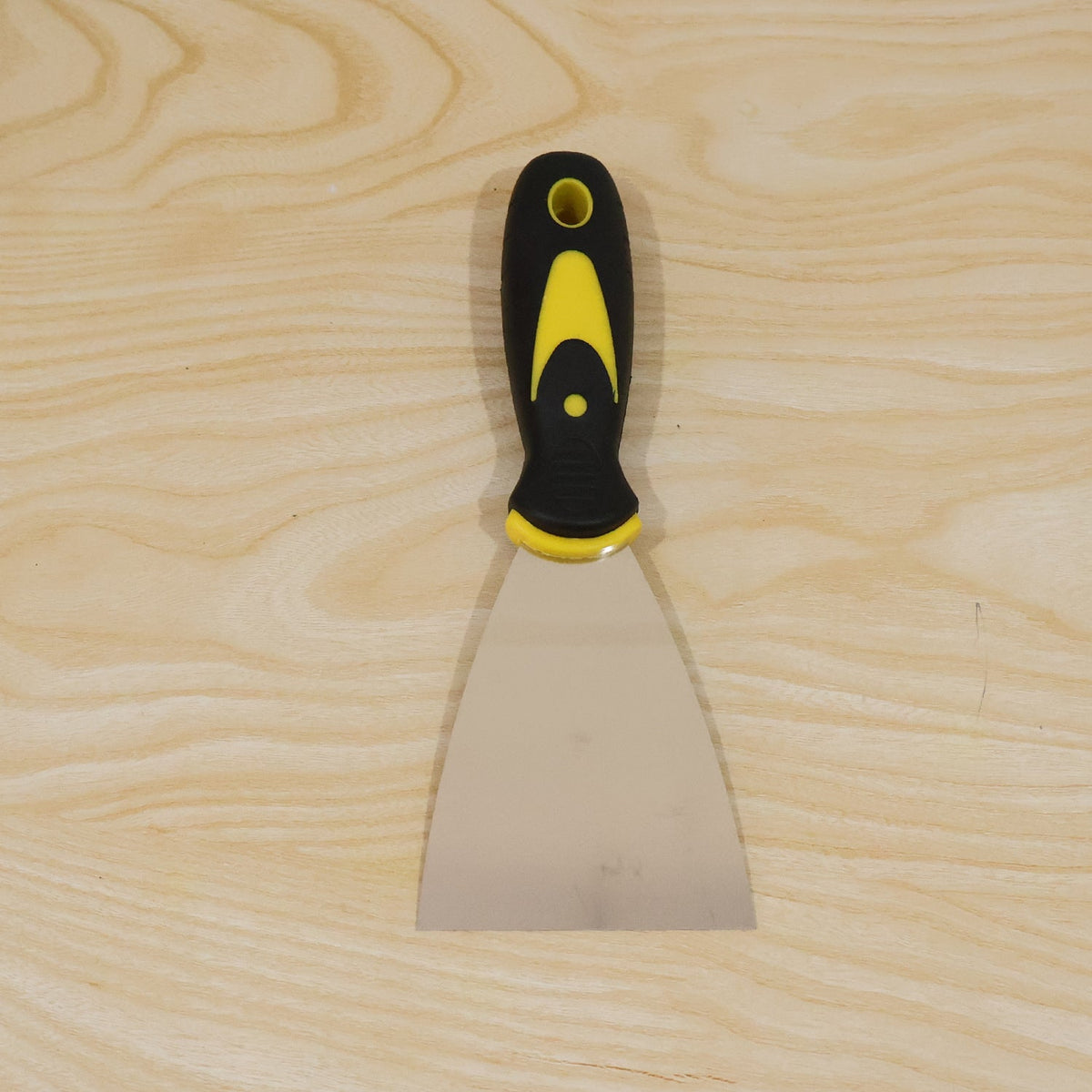 A stainless steel putty knife with a black and yellow handle lies flat on a light-colored wooden surface.