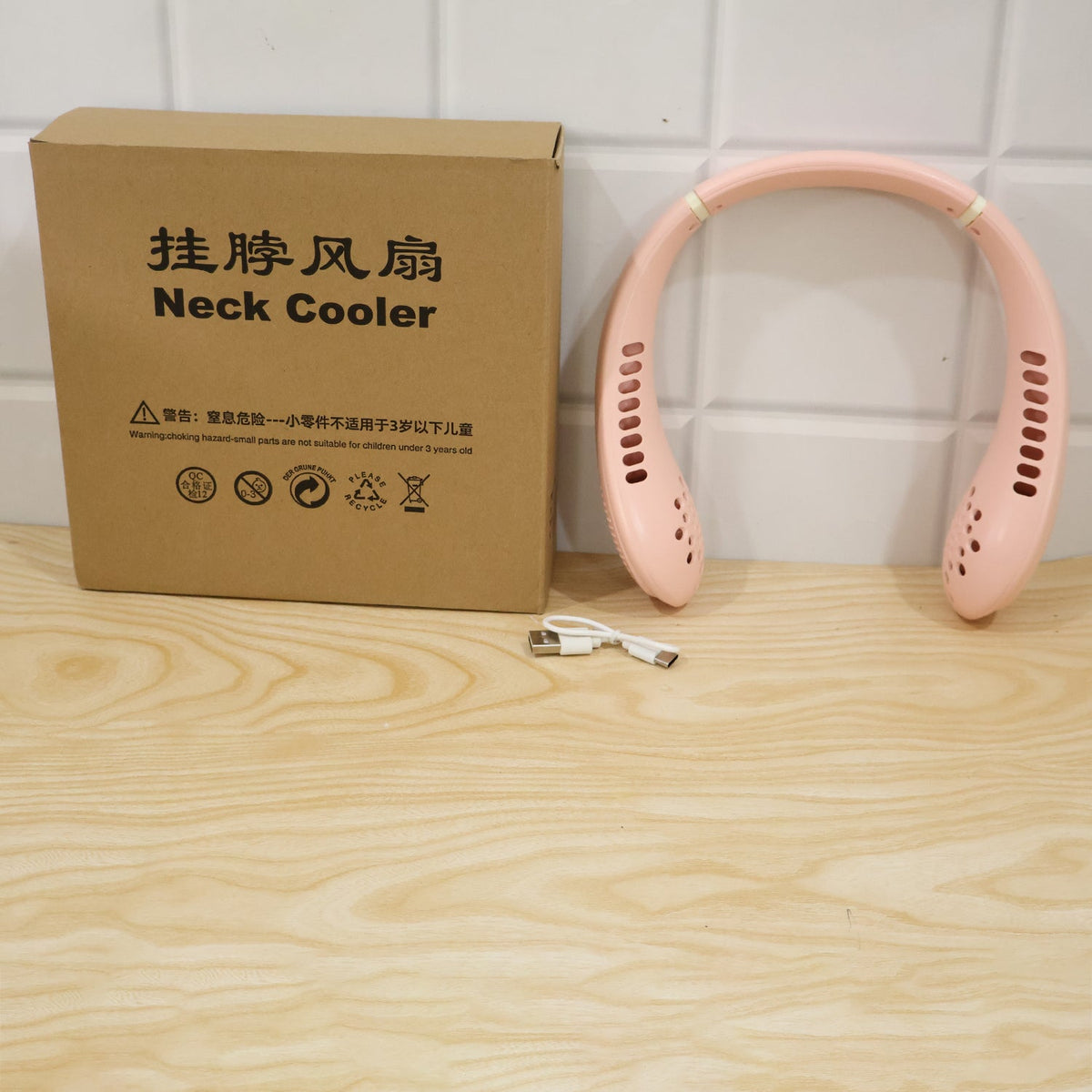 A pink portable neck fan, its brown packaging box, and a white USB cable on a wooden surface.