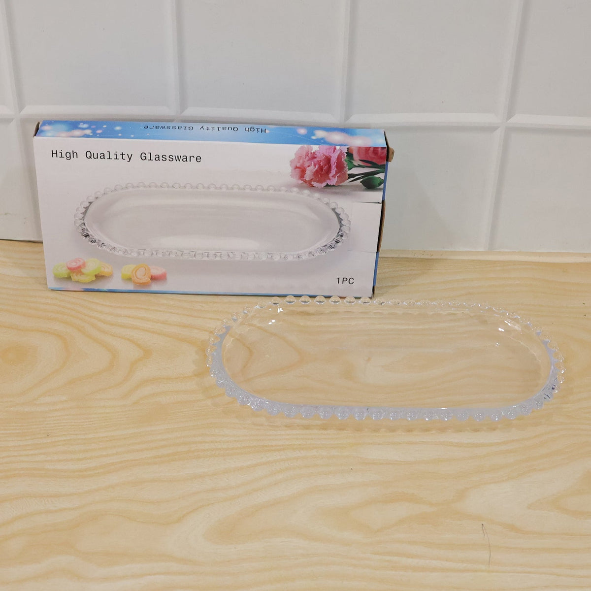 A clear oval glass serving tray is displayed next to its retail packaging on a wooden surface.