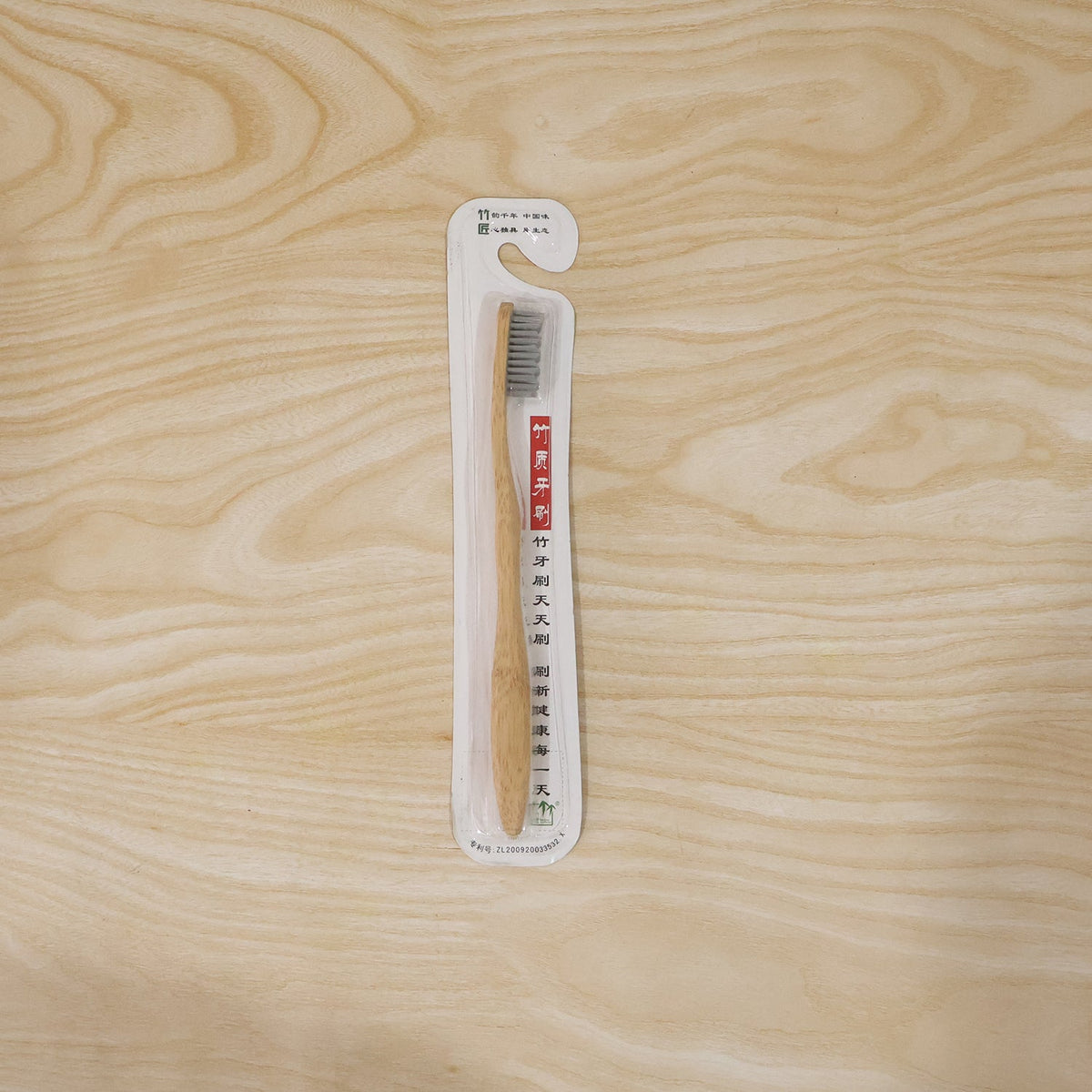 A single bamboo toothbrush with grey bristles is sealed in clear plastic packaging with Chinese text, lying on a light