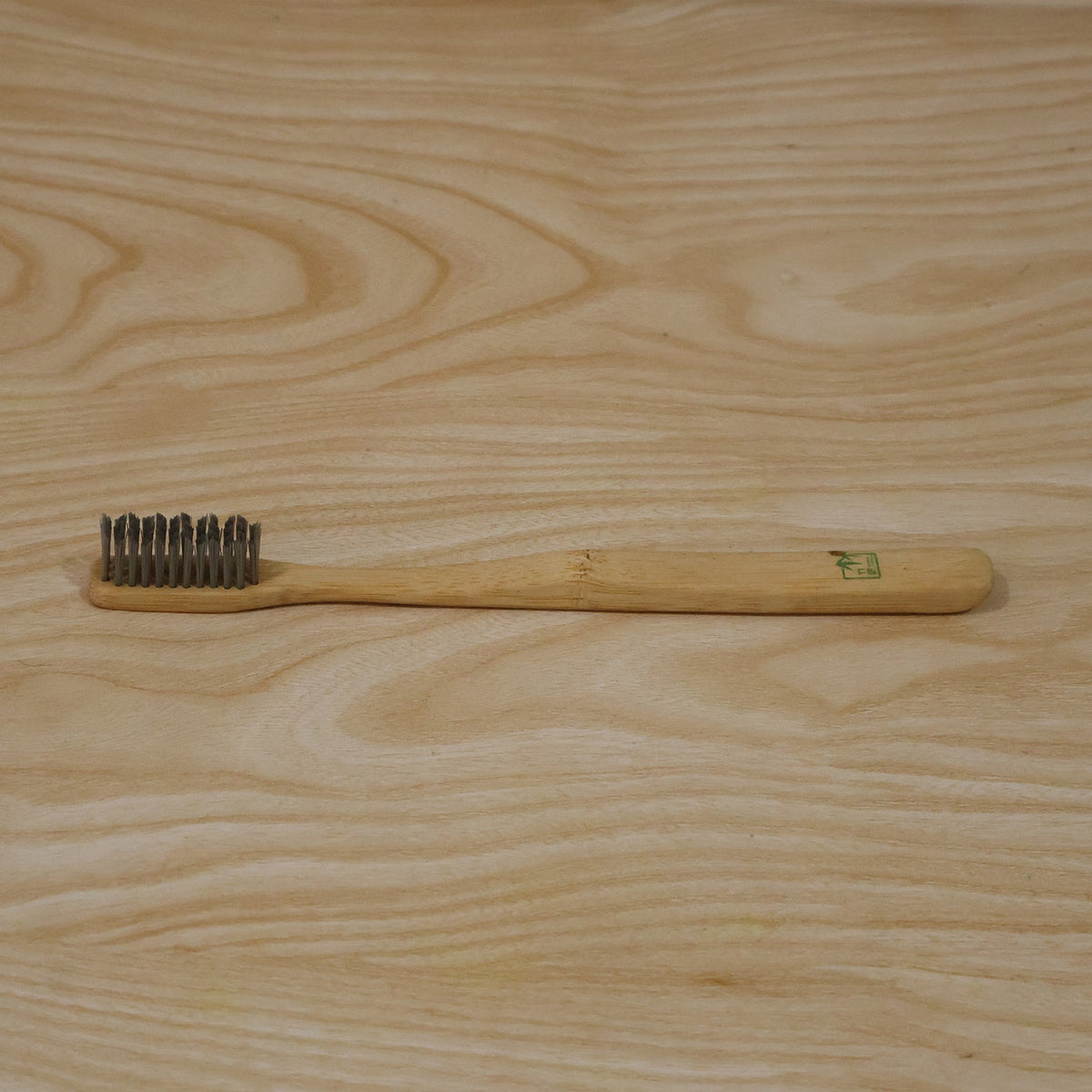 A bamboo toothbrush with grey bristles lies horizontally on a light wooden surface.