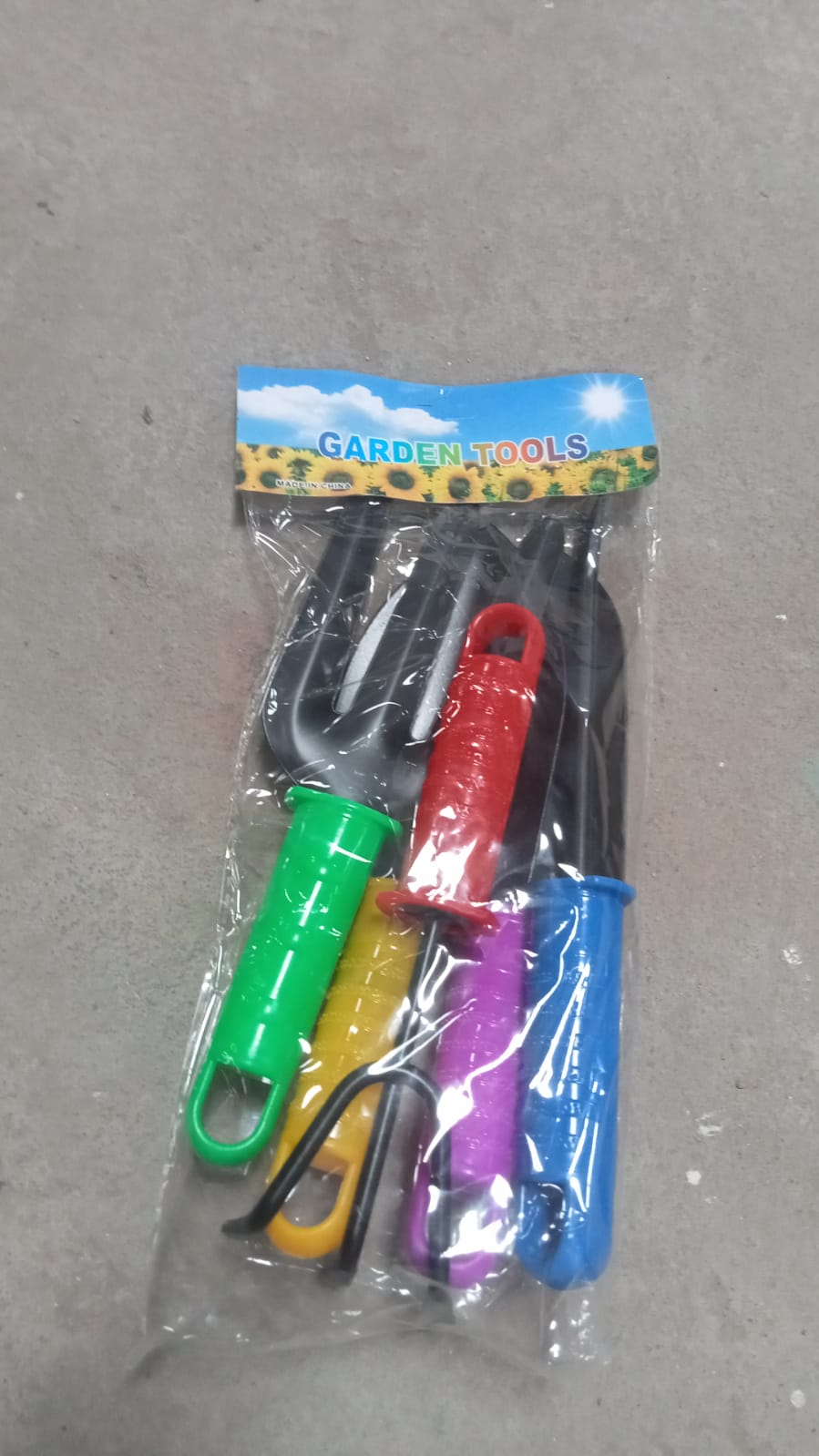 A clear plastic package containing five colorful mini gardening tools is laid on a gray concrete surface.