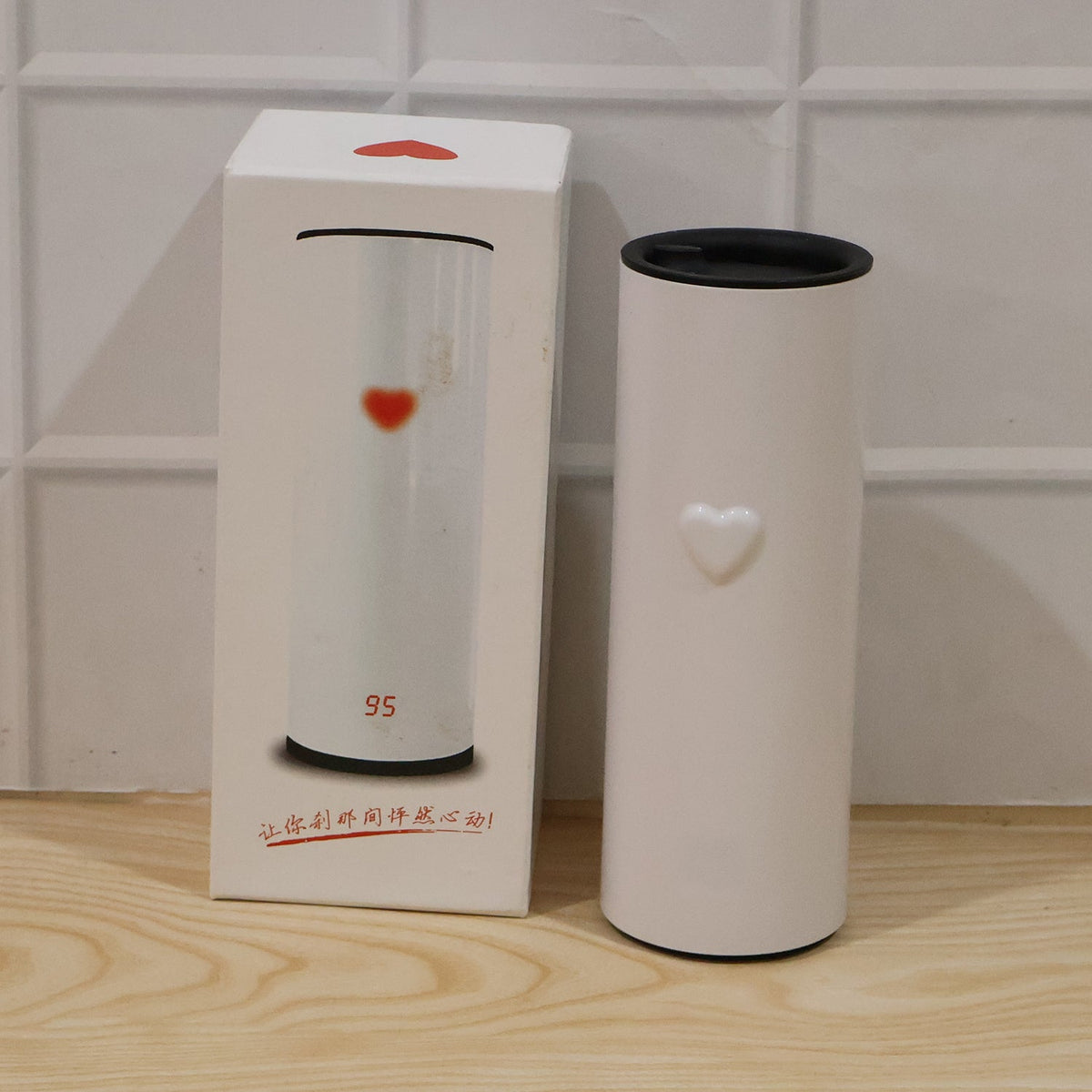 A white thermal tumbler with a heart detail stands next to its product box, which also features a heart design.