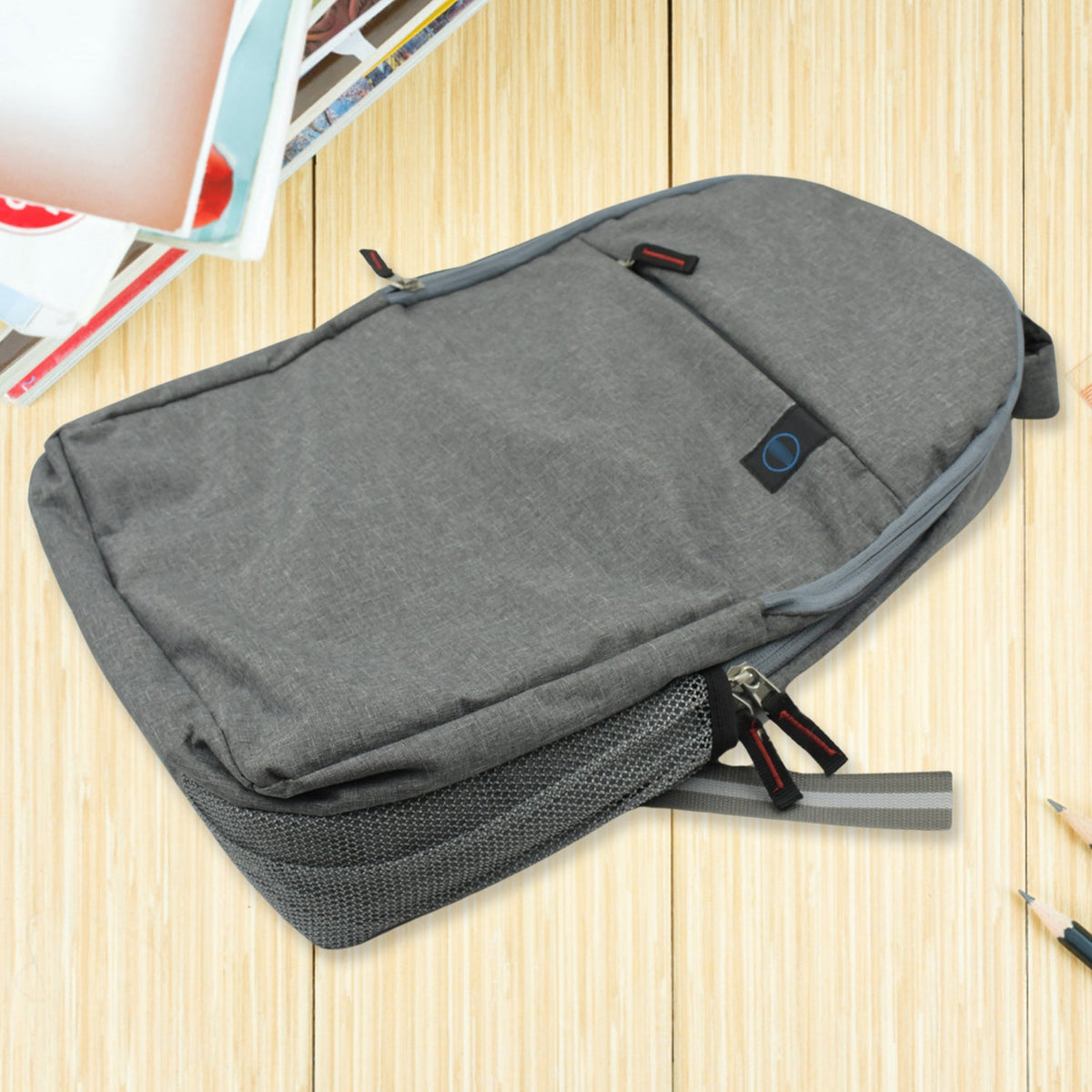A grey backpack with a mesh side pocket and red-accented zippers rests on a wooden surface with pencils.