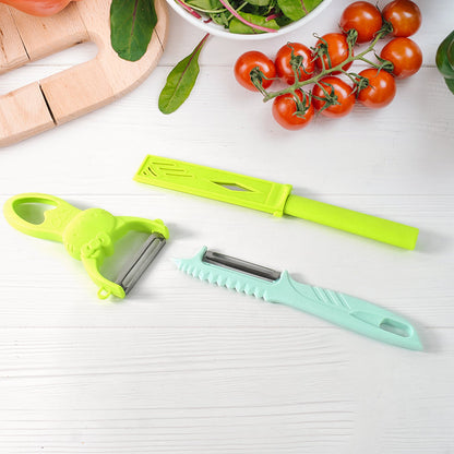 A green peeler, a teal peeler, and a green knife on a white wooden surface, surrounded by fresh vegetables.
