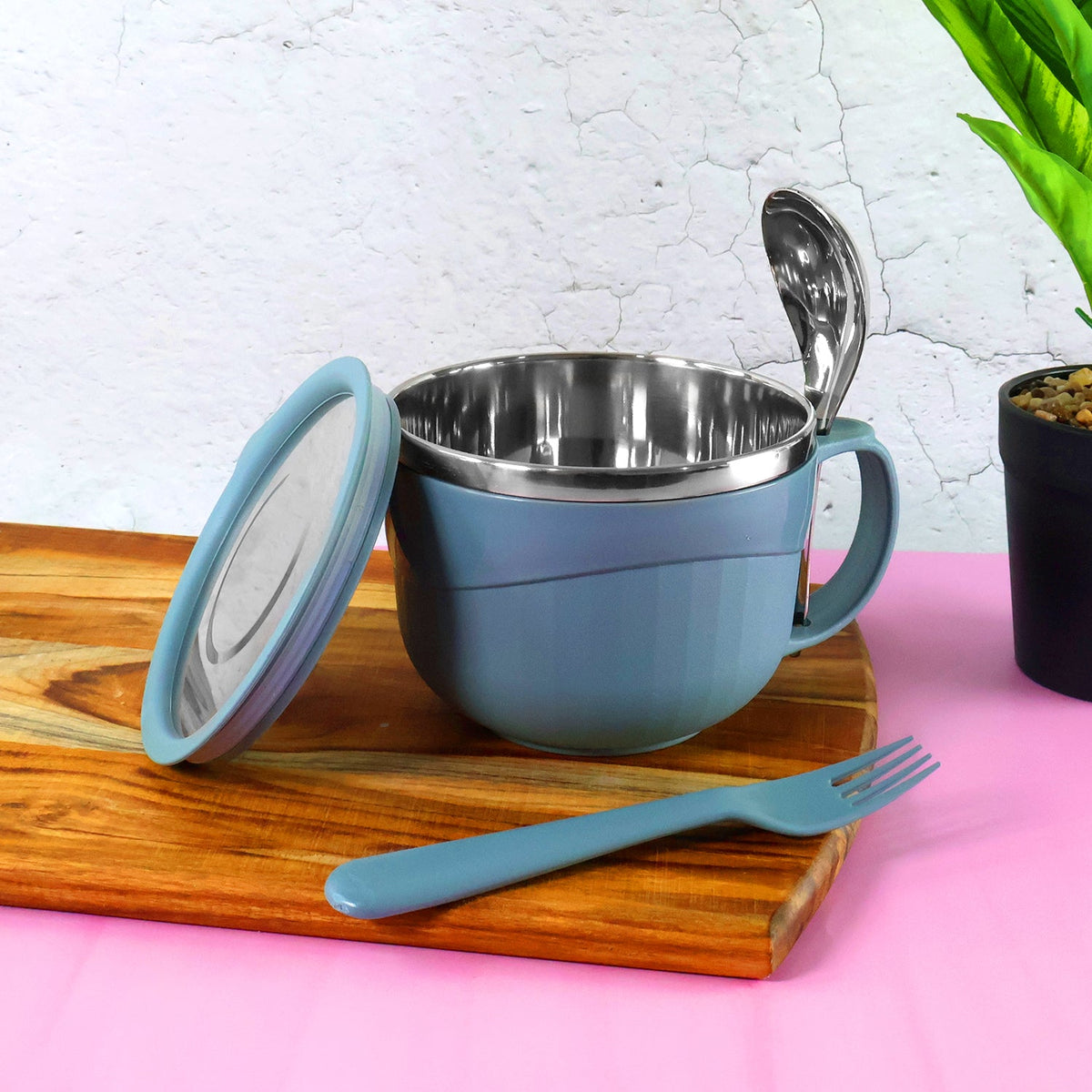 A blue insulated bowl with a lid, a spoon, and a fork on a wooden board against a pink and white background.