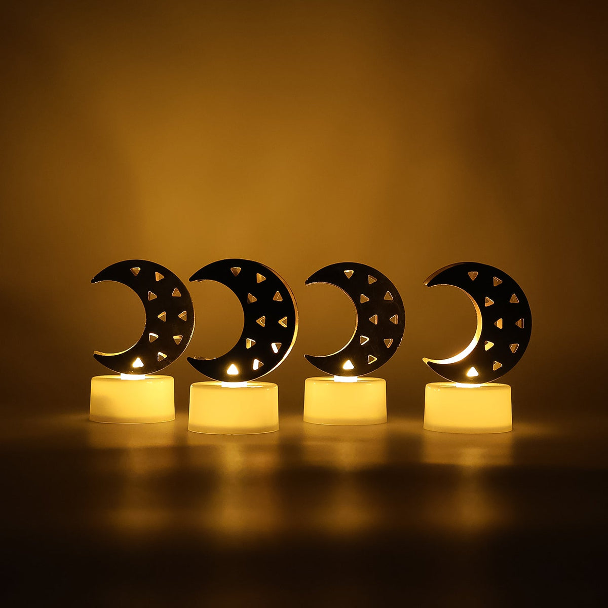 Four crescent moon LED tea lights with warm yellow glow illuminating a dark, plain background.
