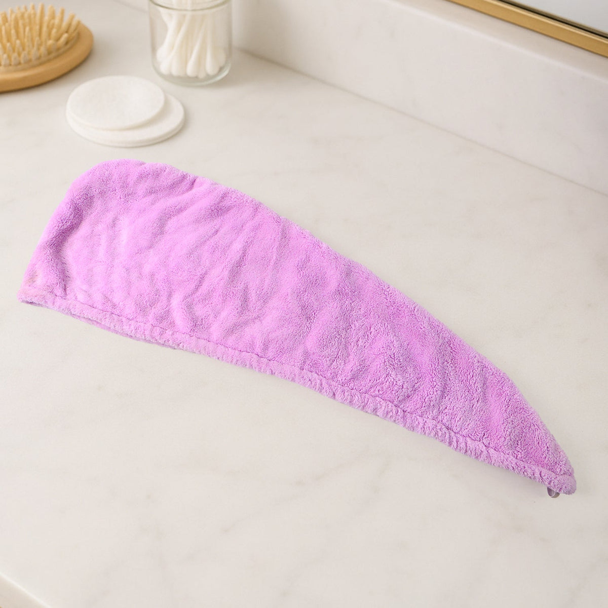 A purple microfiber hair drying wrap towel is laid flat on a white marble countertop next to a brush and cotton swabs.