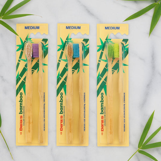 Three twin packs of bamboo toothbrushes with purple, blue, and green bristles on a marble surface with bamboo leaves.