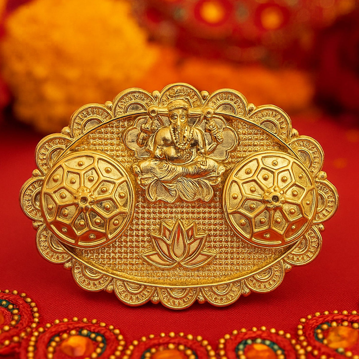 A close-up of a gold-finished chopra featuring an embossed Ganesh, two ornate round containers, and a lotus design on a red