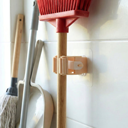 A red broom is held securely by the peach-colored wall-mounted holder on a white tiled wall, with other cleaning tools