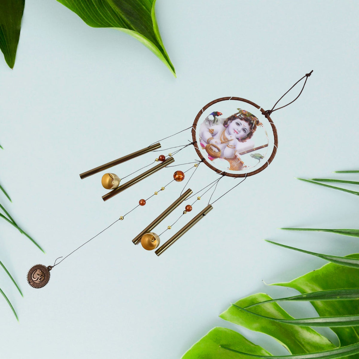 The Krishna wind chime hangs diagonally, surrounded by green leaves on a light blue background.