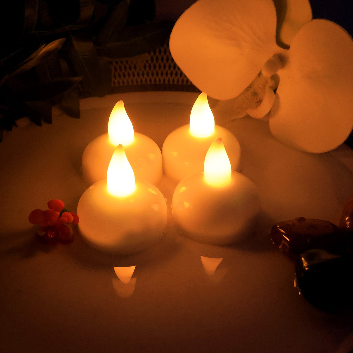 Four white LED floating candles with warm yellow flames on a decorative plate with a white orchid and red berries.