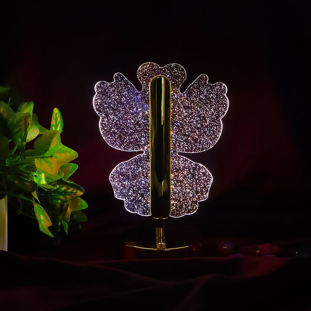 A glowing crystal butterfly-shaped table lamp with a gold base and stem, against a dark background with green foliage.