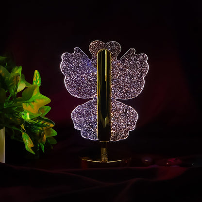 A glowing crystal butterfly-shaped table lamp with a gold base and stem, against a dark background with green foliage.