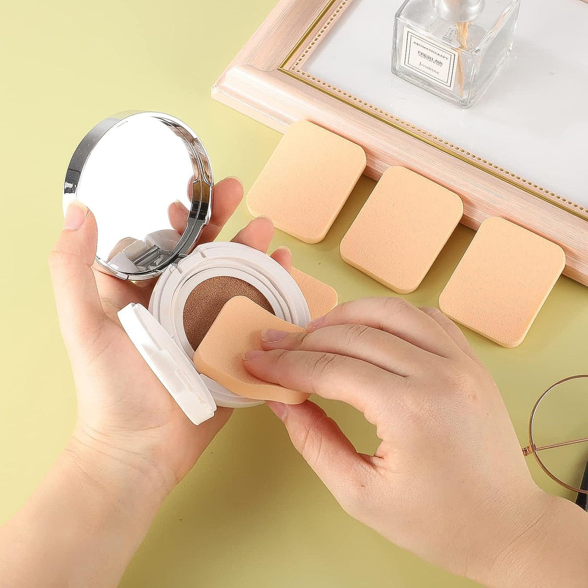 Hands hold a compact cushion foundation with a beige square sponge, with three additional sponges on a light green surface.