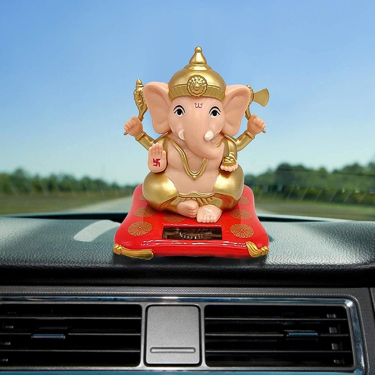 A Ganesha idol on a red base sits on a car dashboard with air vents visible, against a clear sky.