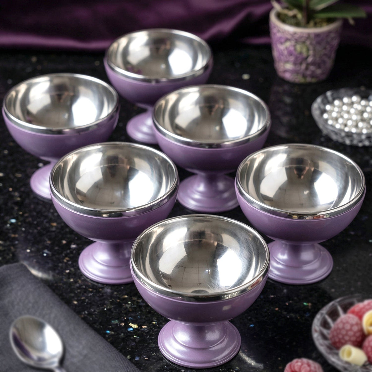 Six matching purple and silver dessert bowls are arranged on a dark, reflective surface next to decorative items.
