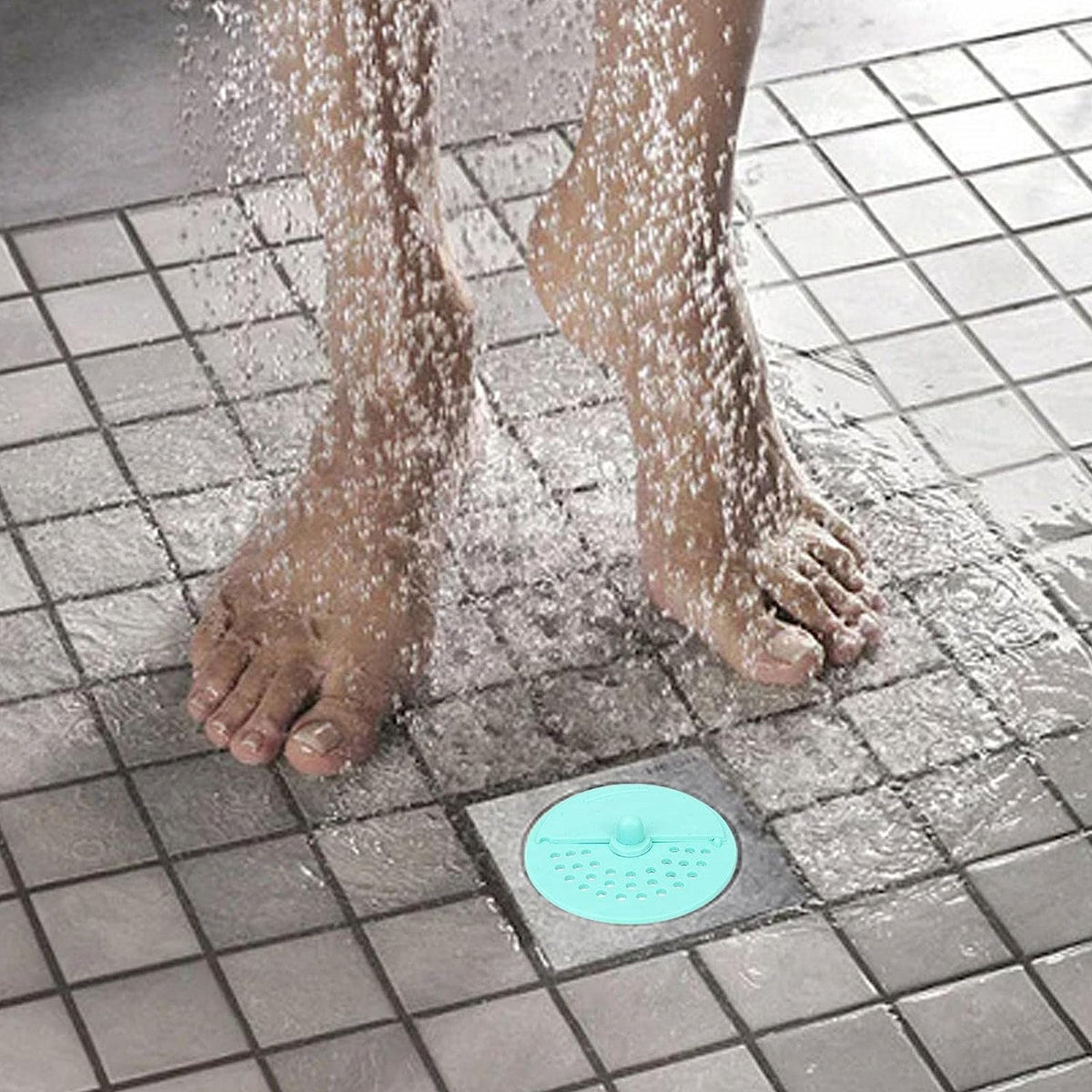 A light blue silicone drain strainer is on a tiled shower floor as water splashes around bare feet.