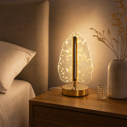 A golden crystal leaf LED table lamp on a wooden nightstand next to a bed, with a small glass votive.