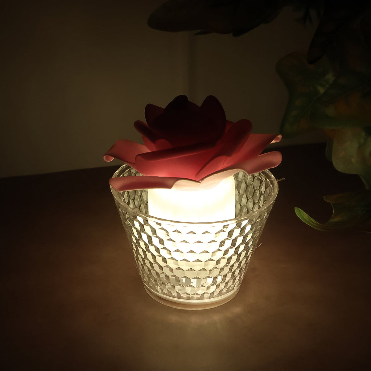 A single red rose-shaped LED candle in a clear textured holder, glowing brightly in a dimly lit room.