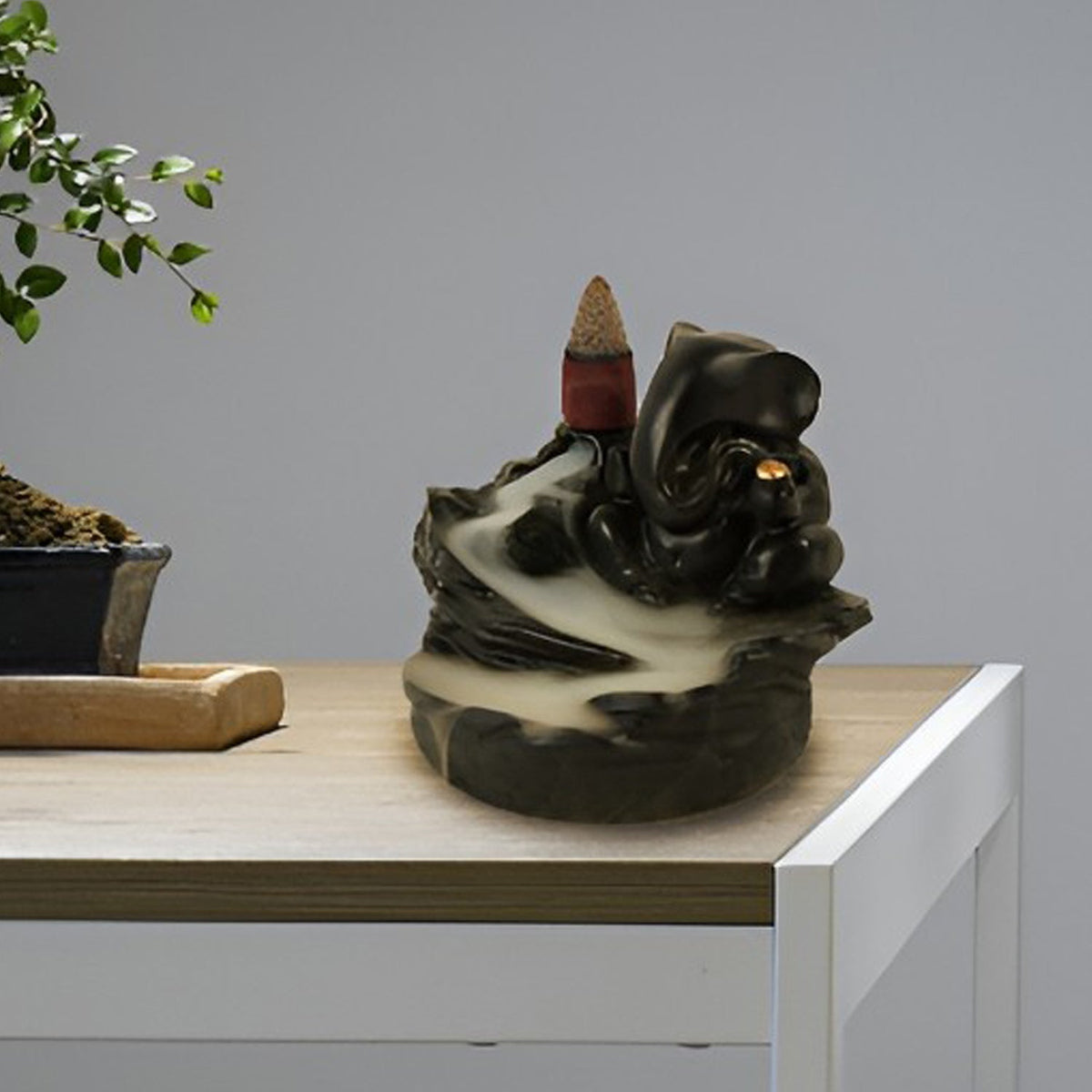 Black Ganesha smoke fountain with a lit incense cone and smoke flowing down, placed on a wooden table next to a bonsai tree.
