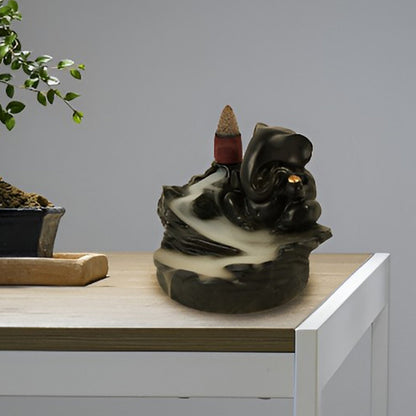 Black Ganesha smoke fountain with a lit incense cone and smoke flowing down, placed on a wooden table next to a bonsai tree.