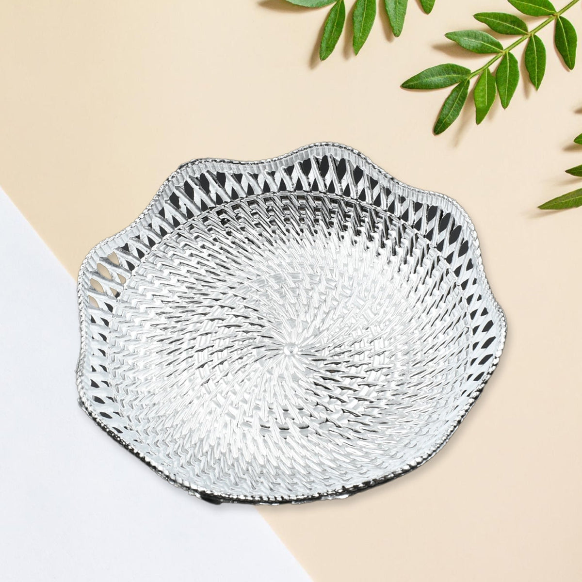A silver plastic scalloped serving bowl with a textured bottom sits on a two-toned beige and white surface with green leaves