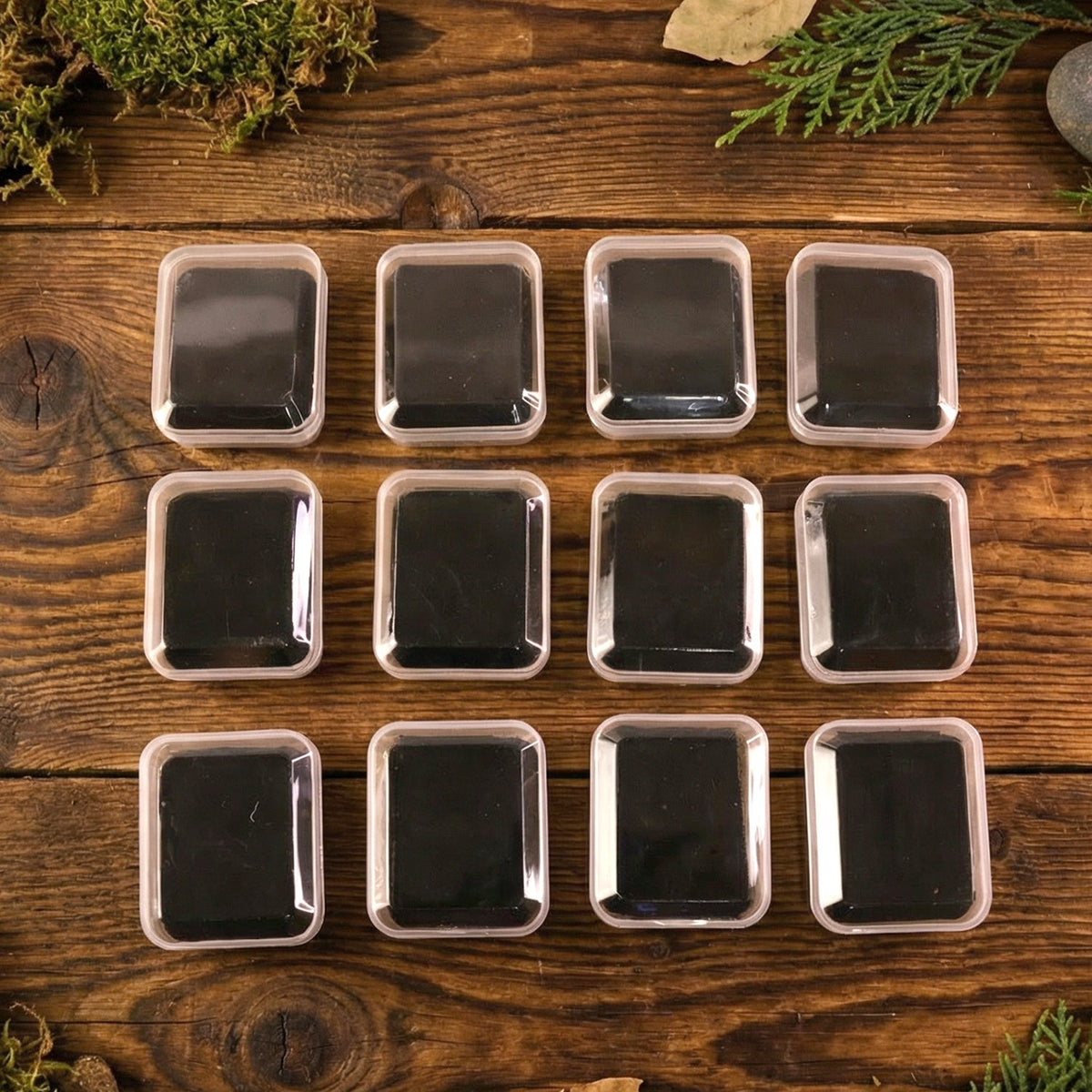 Twelve clear-lidded white jewellery boxes with black inserts displayed on a wooden surface with subtle green and brown