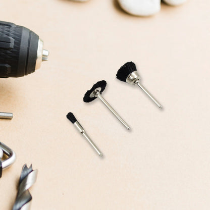 Three black nylon brushes with silver shafts are arranged on a light brown surface next to a drill and other tools.
