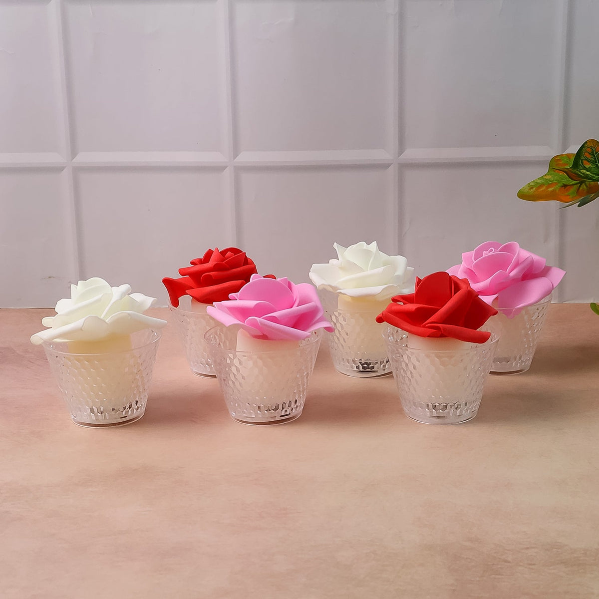 Six rose-shaped LED candles in clear textured holders, unlit, arranged on a light pink surface with a tiled wall.