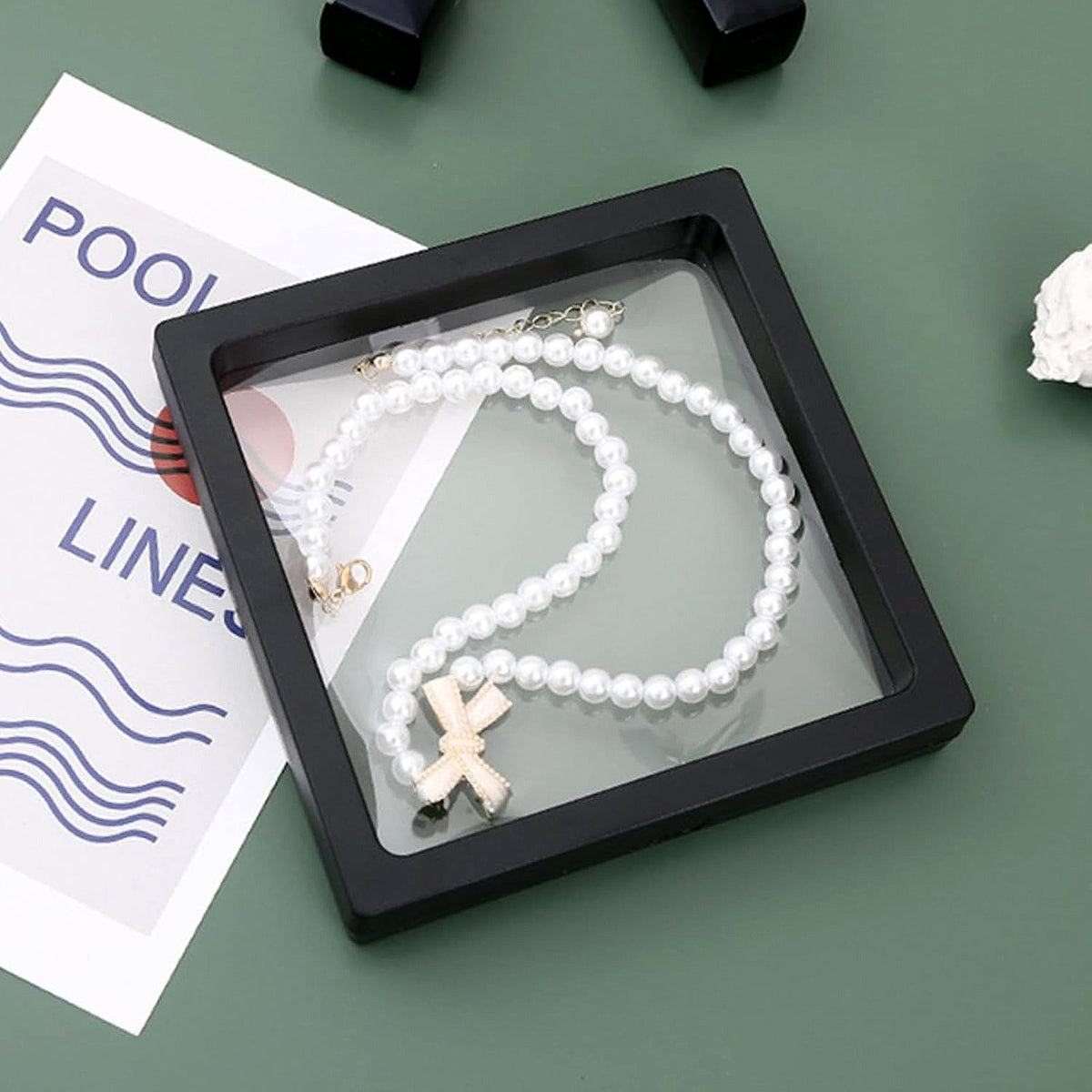 Black square display box with a pearl necklace and bow charm floating inside on a green background.