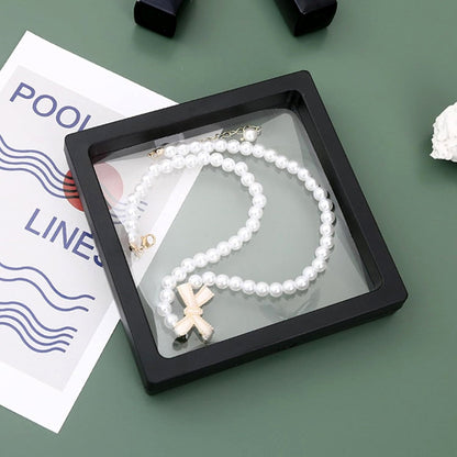 Black square display box with a pearl necklace and bow charm floating inside on a green background.